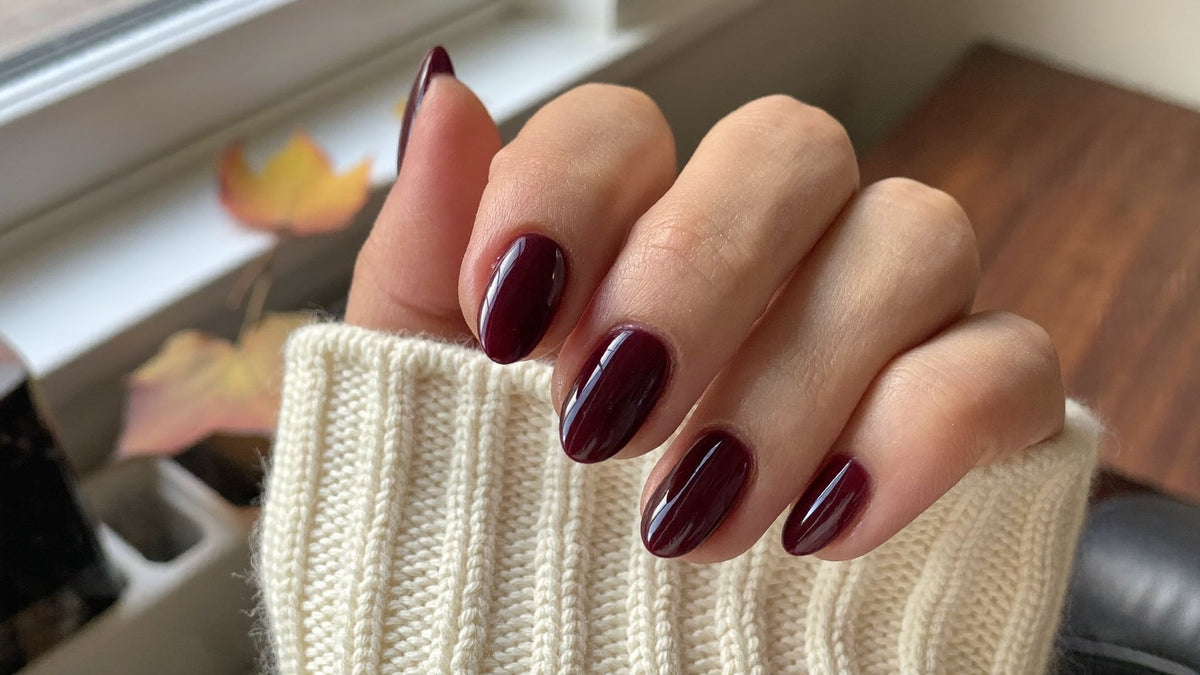 Autumn nails 2026: hand with glossy deep burgundy gel nails holding a cream knit sweater cuff by a window with autumn leaves