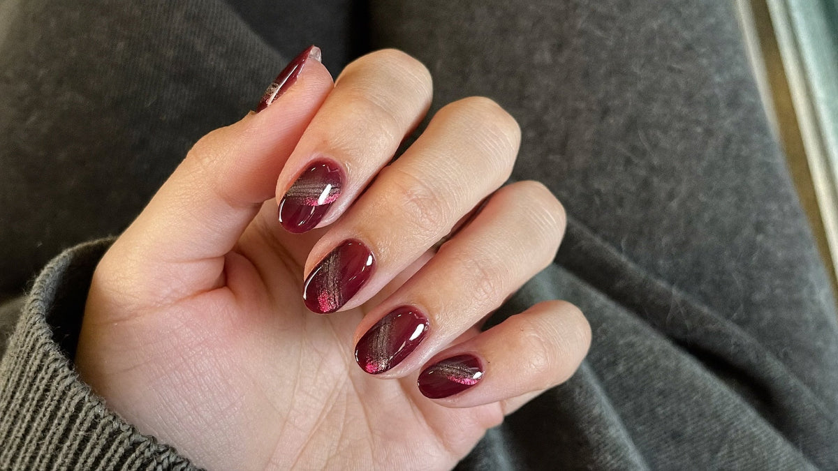 Cat eye nails: hand with burgundy magnetic gel polish showing a diagonal band of shimmering light on each nail, resting on a grey knit sweater